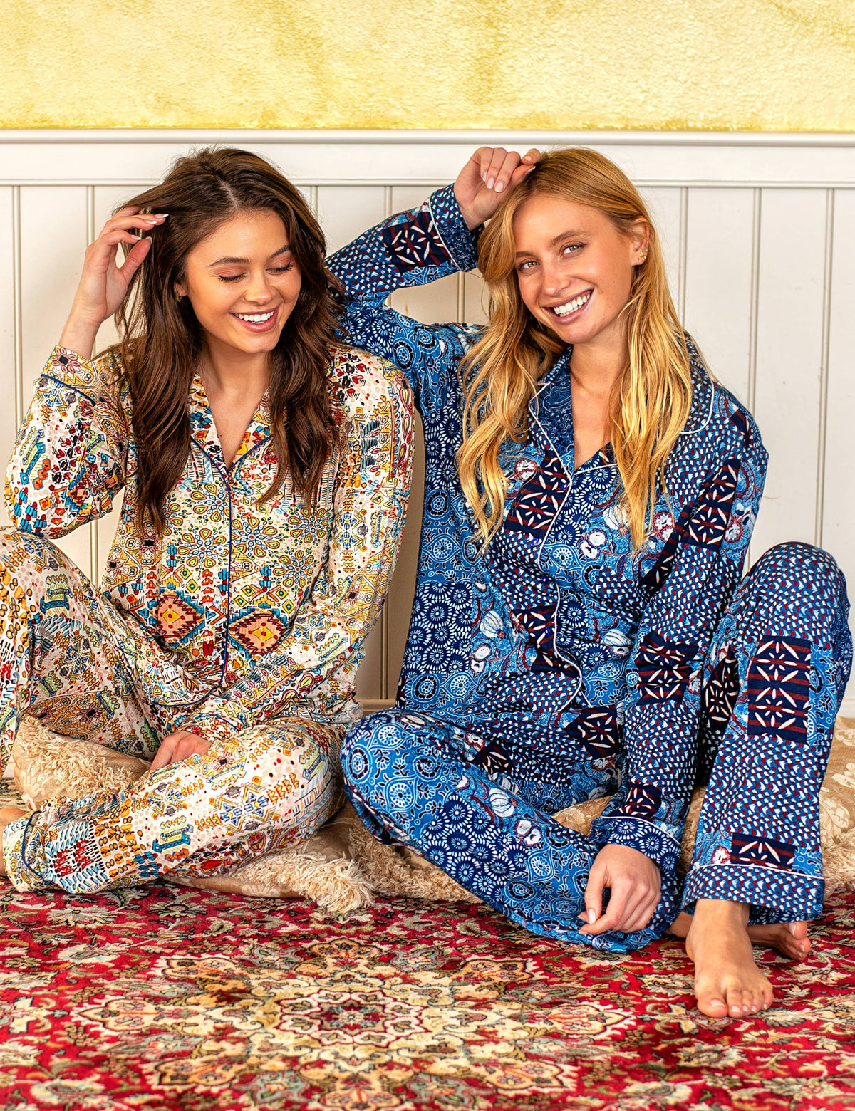 Two women sitting on a patterned rug wearing matching patterned pajamas in a cozy indoor setting. The woman on the left wears a multicolor paisley print pajama set, and the woman on the right wears a blue floral print pajama set.