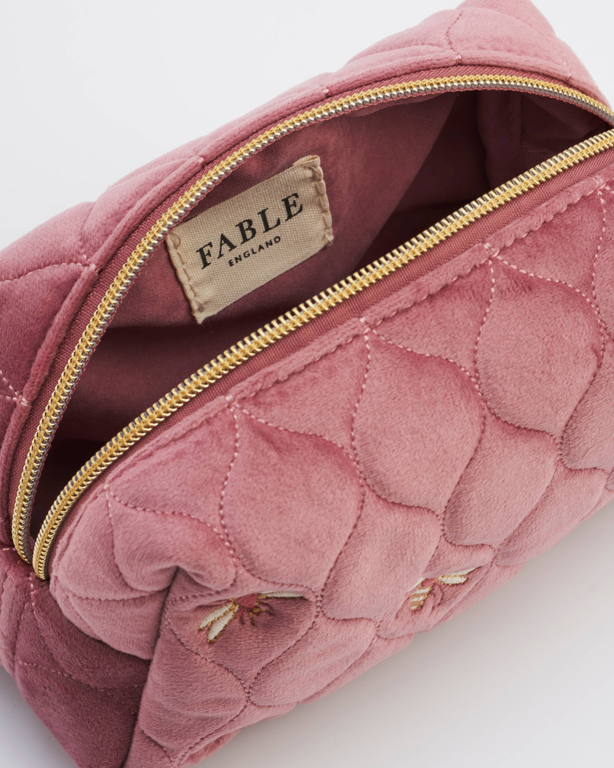 A close-up of a dusty pink quilted cosmetic pouch by FABLE, showing its gold zipper, interior lining, and embroidered bee details on the front.