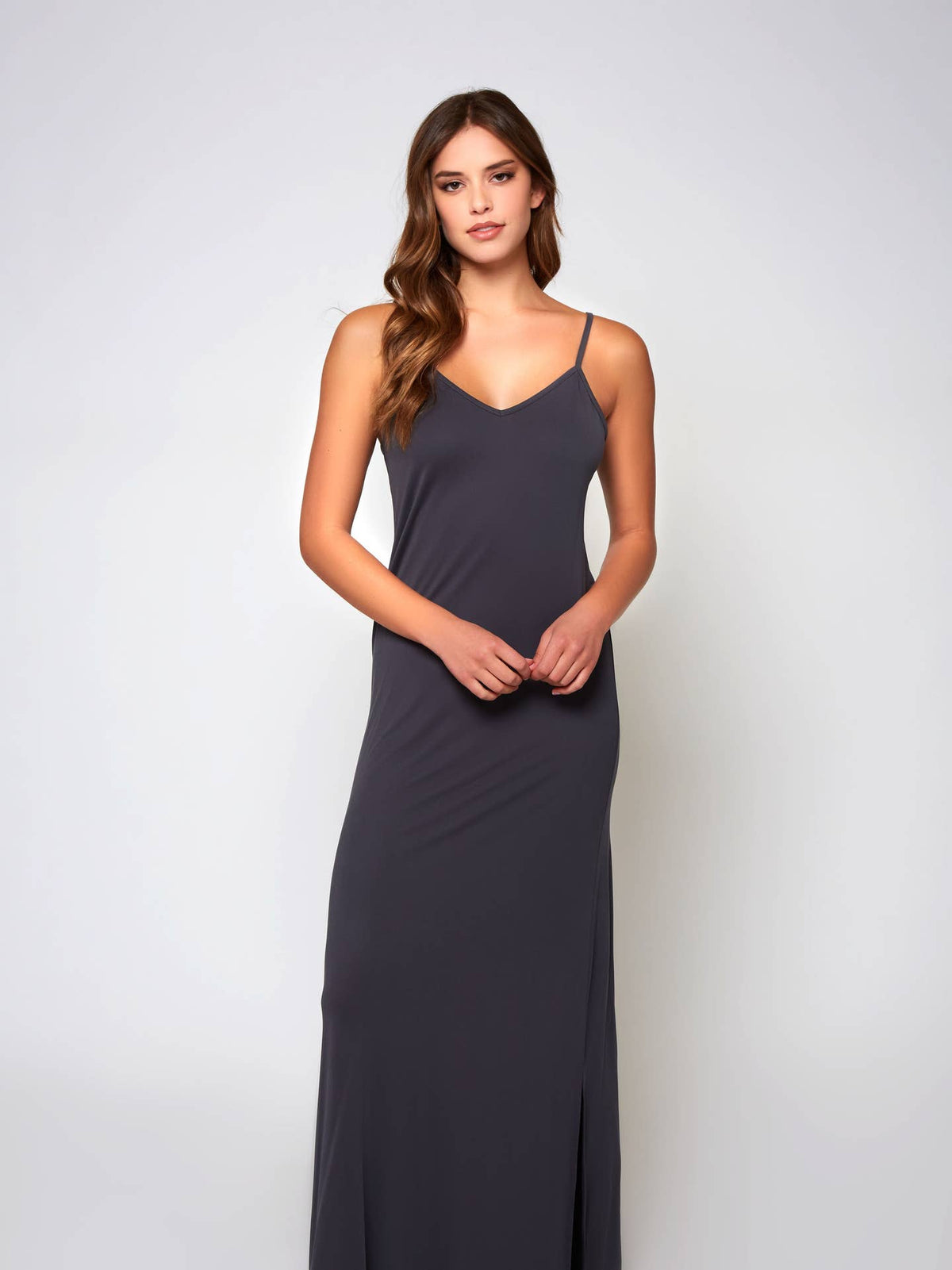 A woman modeling a dark charcoal gray maxi slip dress with thin spaghetti straps and a V-neckline, standing against a light gray studio background.