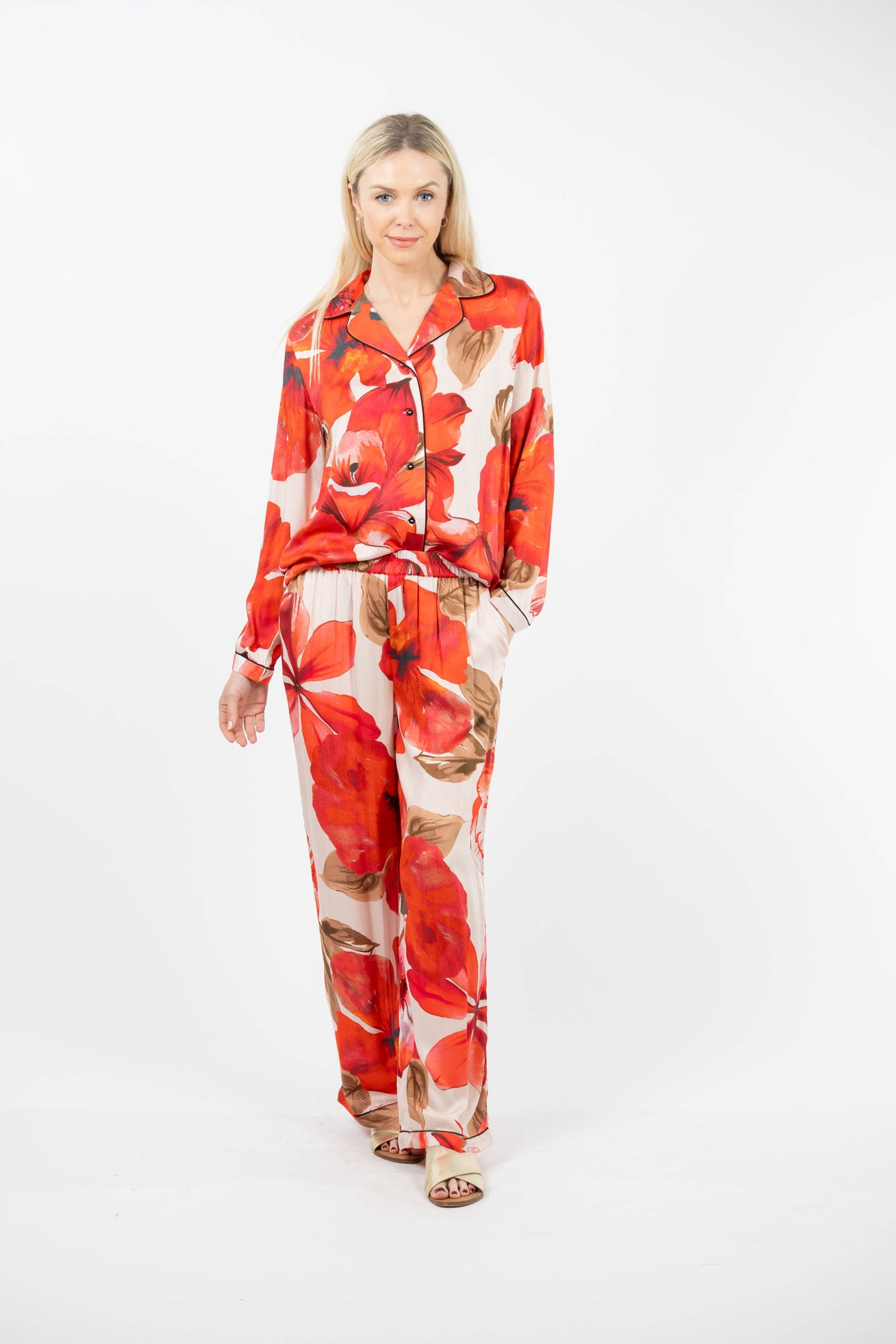 A woman modeling a matching two-piece pajama set with a bold red and orange floral print on a cream background, photographed against a seamless white studio background.