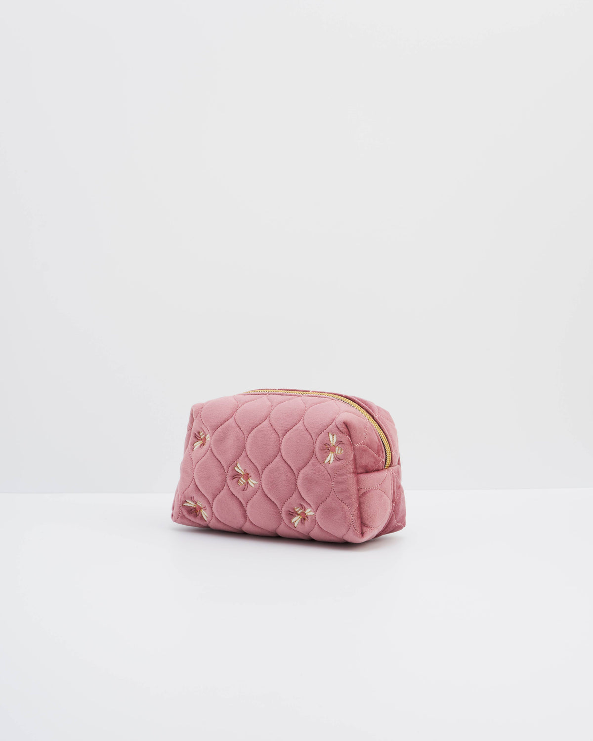 A pink quilted cosmetic pouch with embroidered floral details and a gold zipper, set against a plain white studio background.