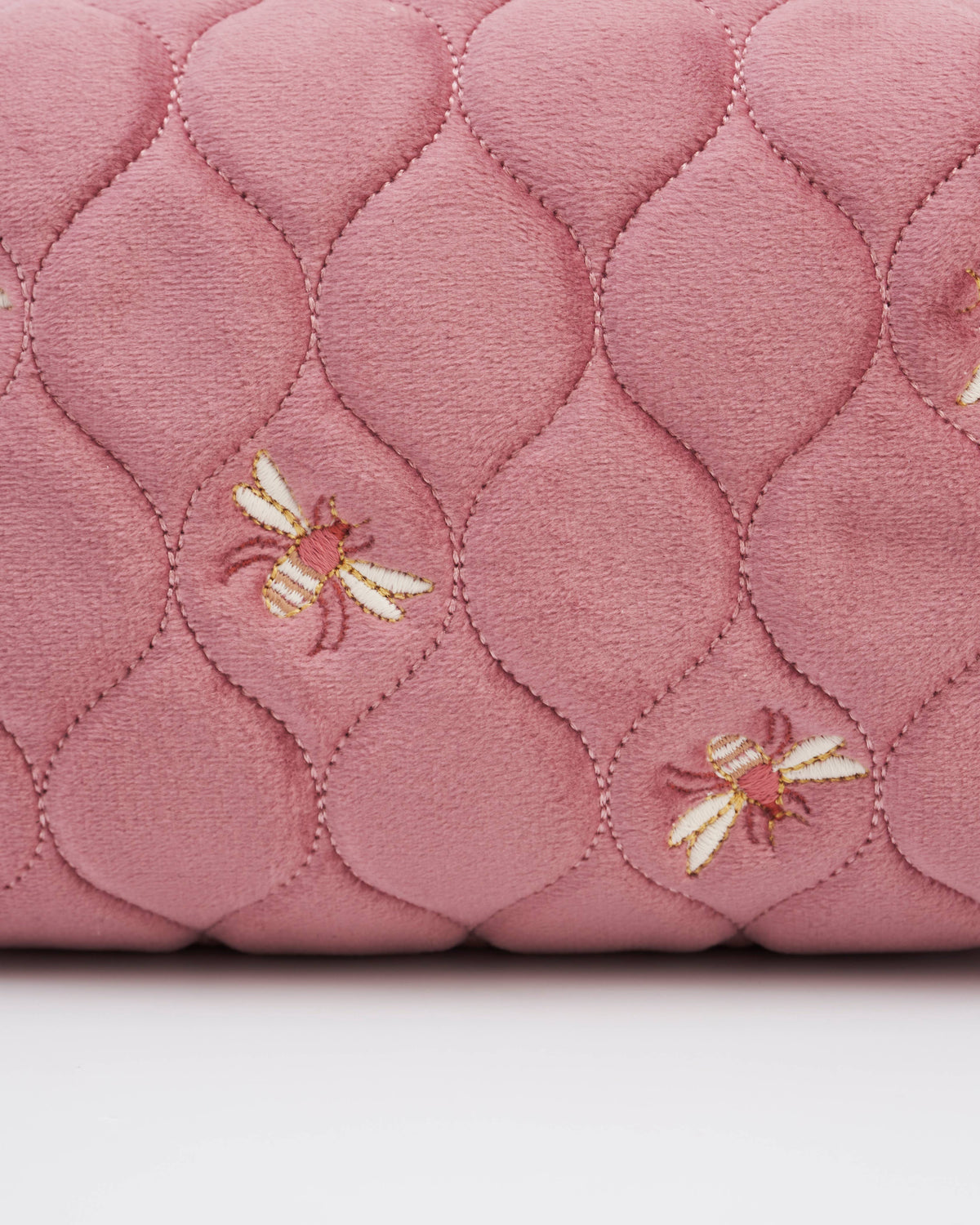 A close-up of a dusty pink quilted fabric with a repeating diamond pattern and small embroidered bees in cream, gold, and brown.