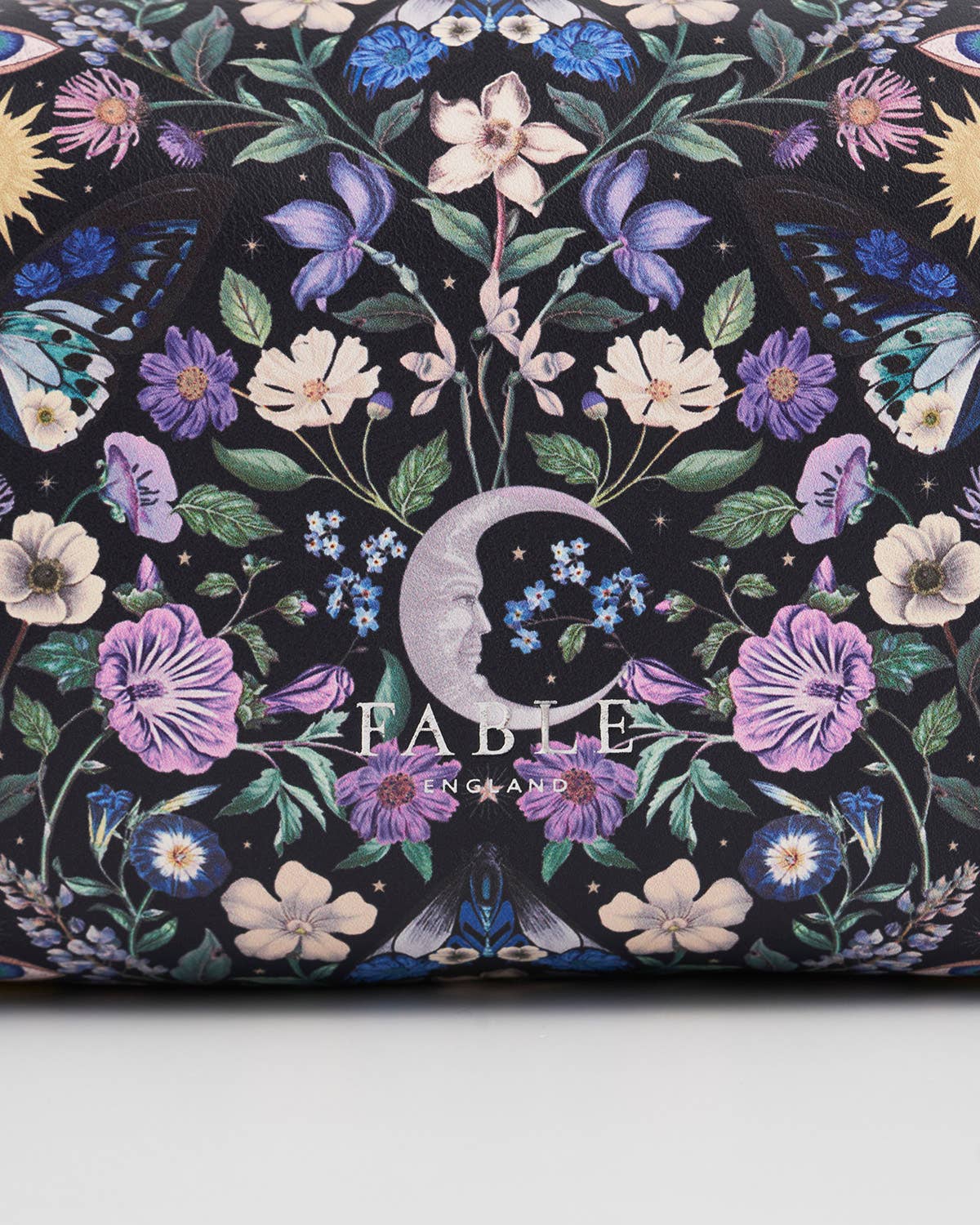 A close-up of a Fable by Gatsby clutch or wallet featuring a dark background covered in a detailed botanical and celestial print with purple flowers, blue butterflies, green leaves, and a crescent moon.