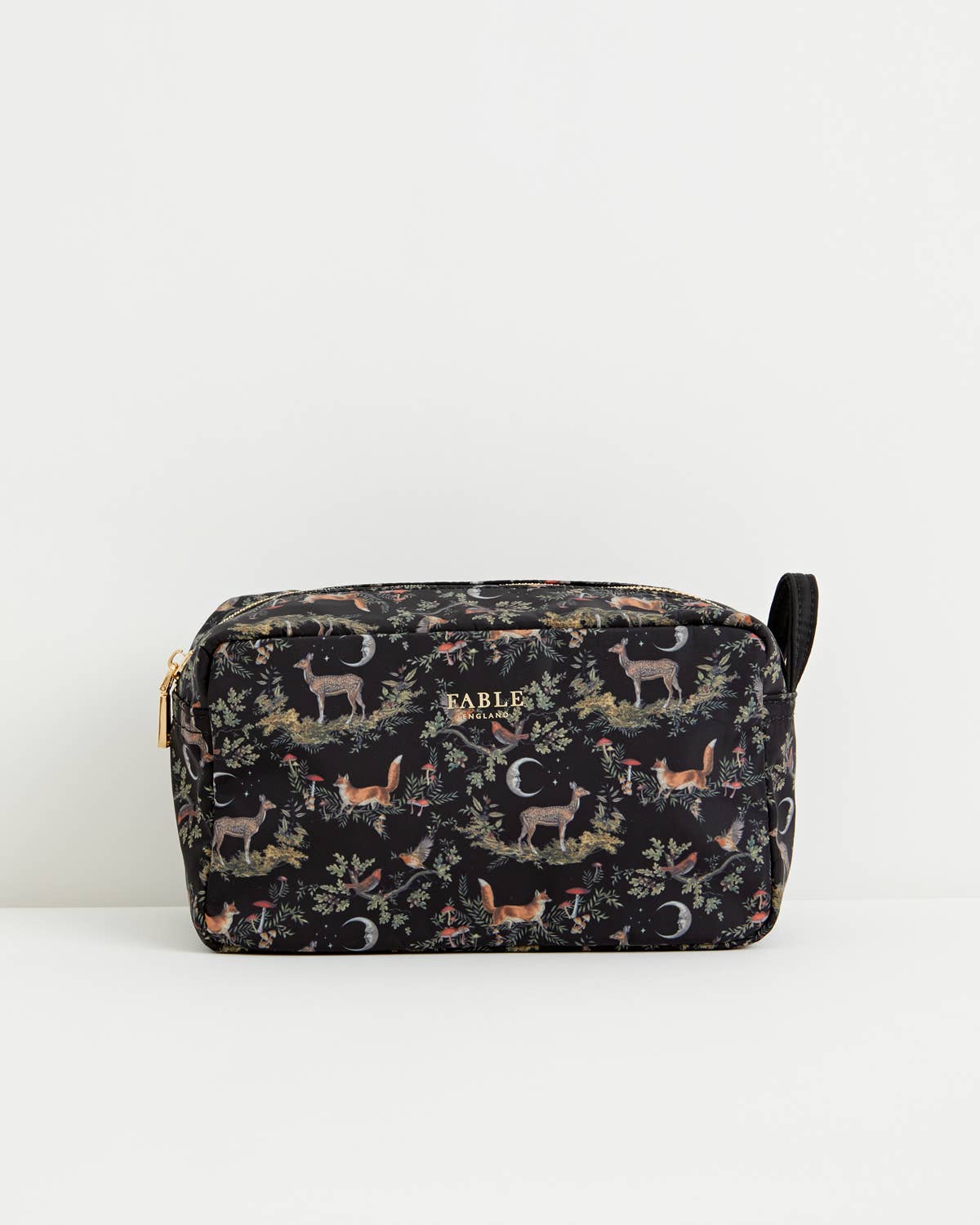 A rectangular cosmetic pouch by Table with a black background and an all-over woodland print featuring deer, foxes, trees, and crescent moons. The pouch has a gold zipper, a small gold Table logo on the front, and a black wrist strap, photographed against a clean white background.
