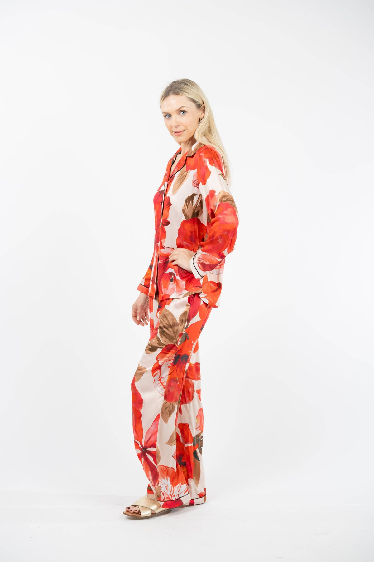 A woman modeling a matching two-piece pajama set with a bold red, orange, and brown floral pattern on a cream background, photographed against a seamless white studio background.
