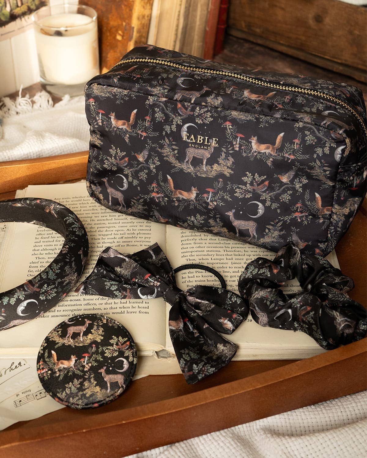 A matching vanity set with a zippered pouch, headband, round compact, bow, and scrunchies in a dark woodland print featuring deer, moths, and crescent moons, styled on an open book in a wooden tray.