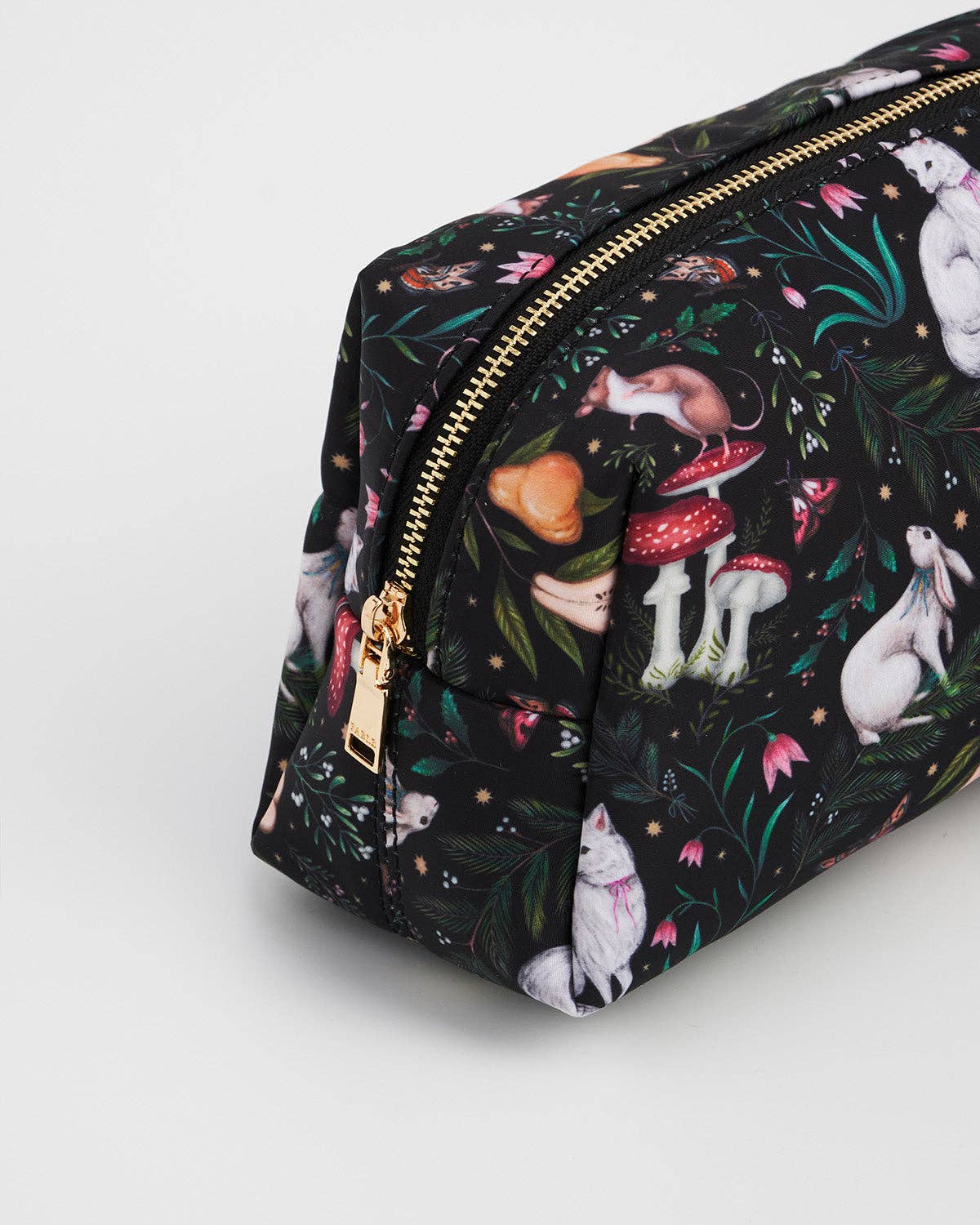 A black zippered cosmetic pouch featuring a whimsical forest and animal print with white rabbits, red-and-white mushrooms, green leaves, and gold-tone hardware, photographed on a light gray background.