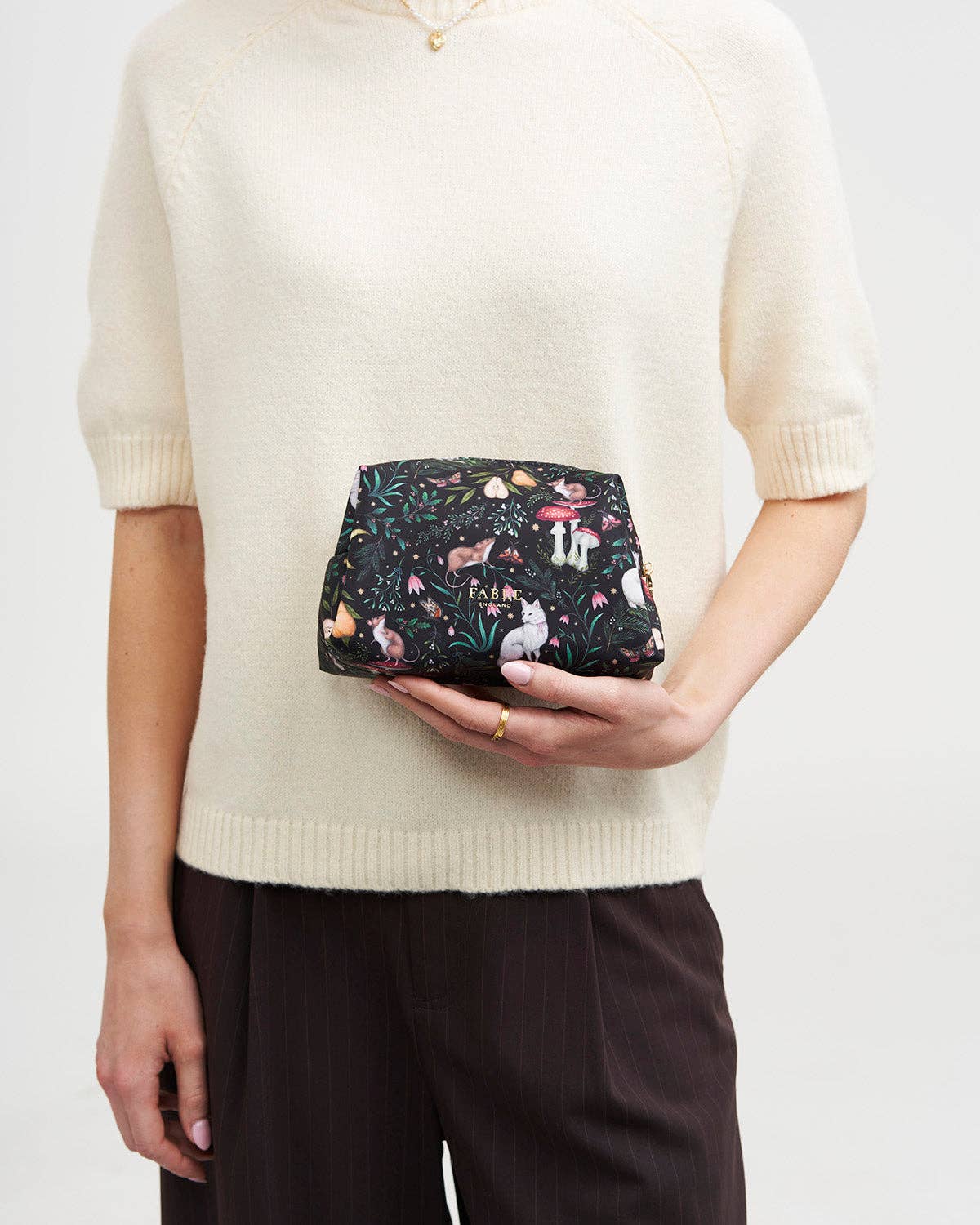 A person wearing a cream knit sweater and dark brown trousers holds a small black clutch bag with a colorful woodland animal print featuring deer, foxes, and mushrooms.