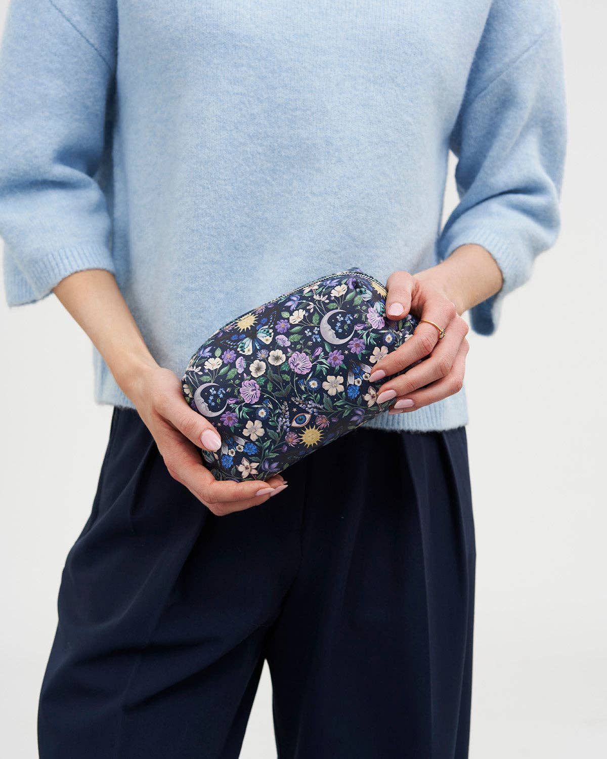 A person wearing a light blue sweater and dark navy trousers holds a small zip pouch with an intricate multicolor floral and paisley pattern against a plain light background.