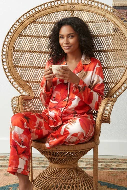 A woman wearing a red and white floral satin pajama set sits in a large woven rattan peacock chair, holding a white mug.