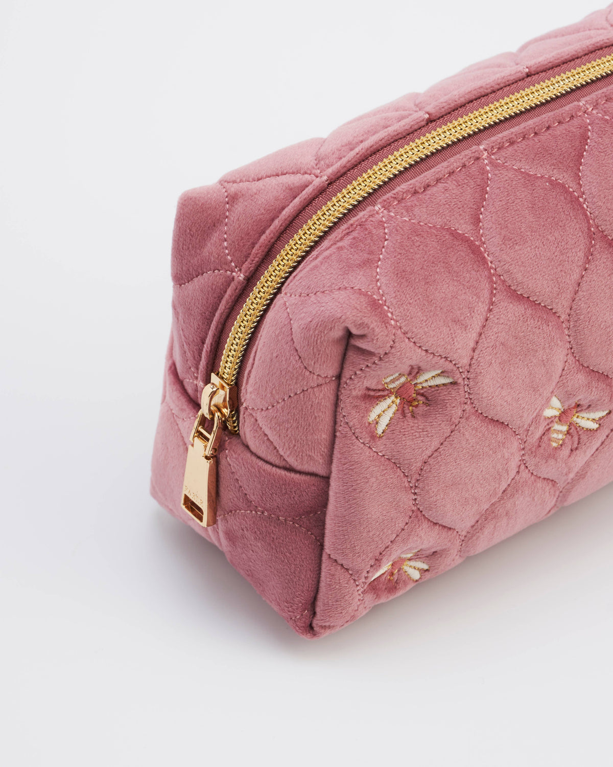 A close-up of a dusty pink quilted suede cosmetic pouch with gold-tone hardware and embroidered floral details, set against a clean white background.