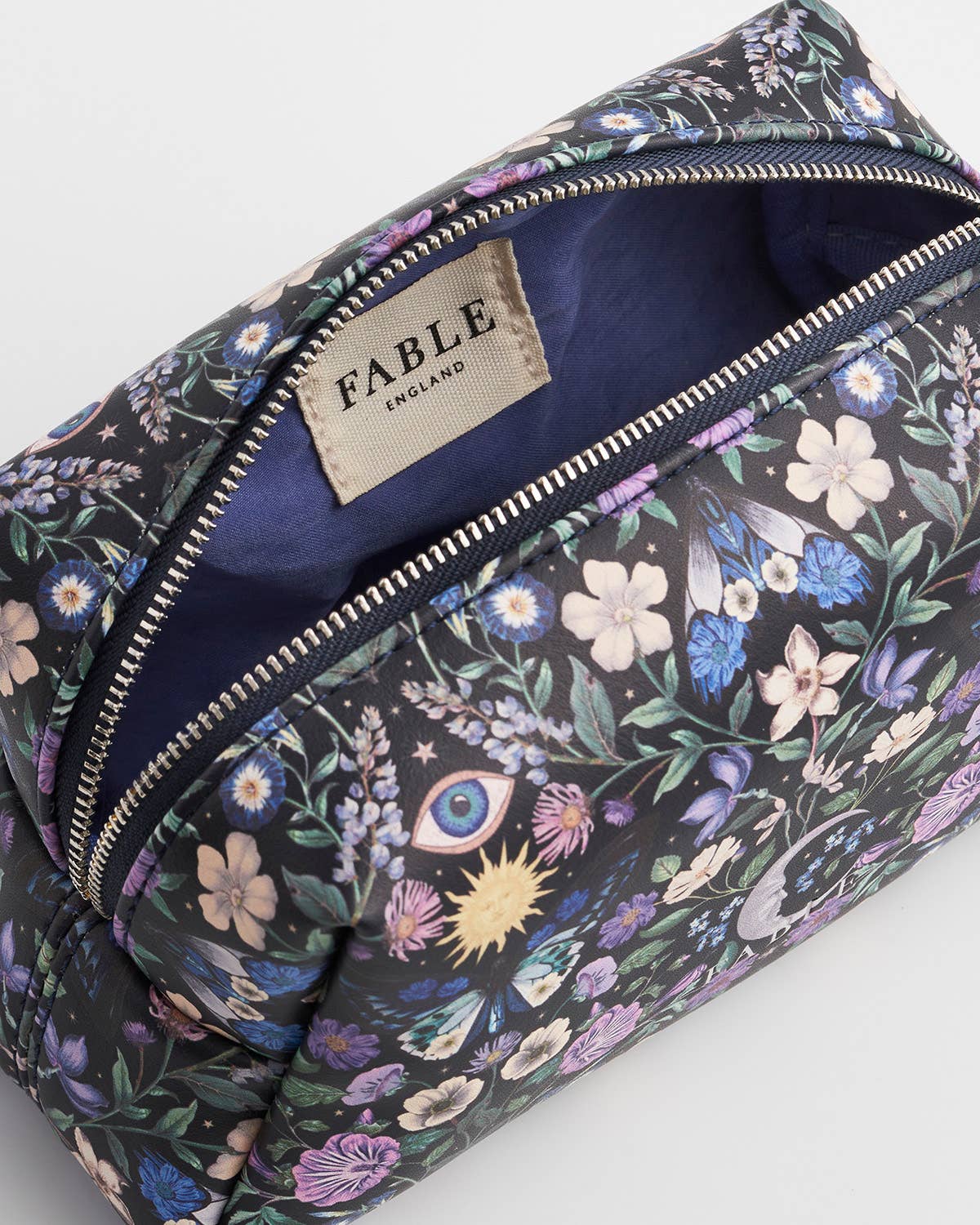 A close-up of a Fable England cosmetic pouch with a dark background and a colorful floral and mystical print featuring an eye, rabbit, sun, and stars. The pouch is shown open, revealing a navy blue interior and a beige brand label.